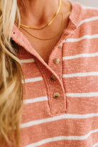 Close-up of a person wearing a ribbed knit henley-style top in a muted peach-pink color with thin white horizontal stripes and brown buttons. A gold chain necklace is also visible.