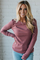 Woman wearing a mauve long-sleeve top and light blue jeans against a white brick wall.