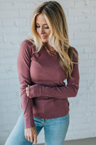 Woman wearing a maroon long-sleeve top and light blue jeans against a white brick wall.
