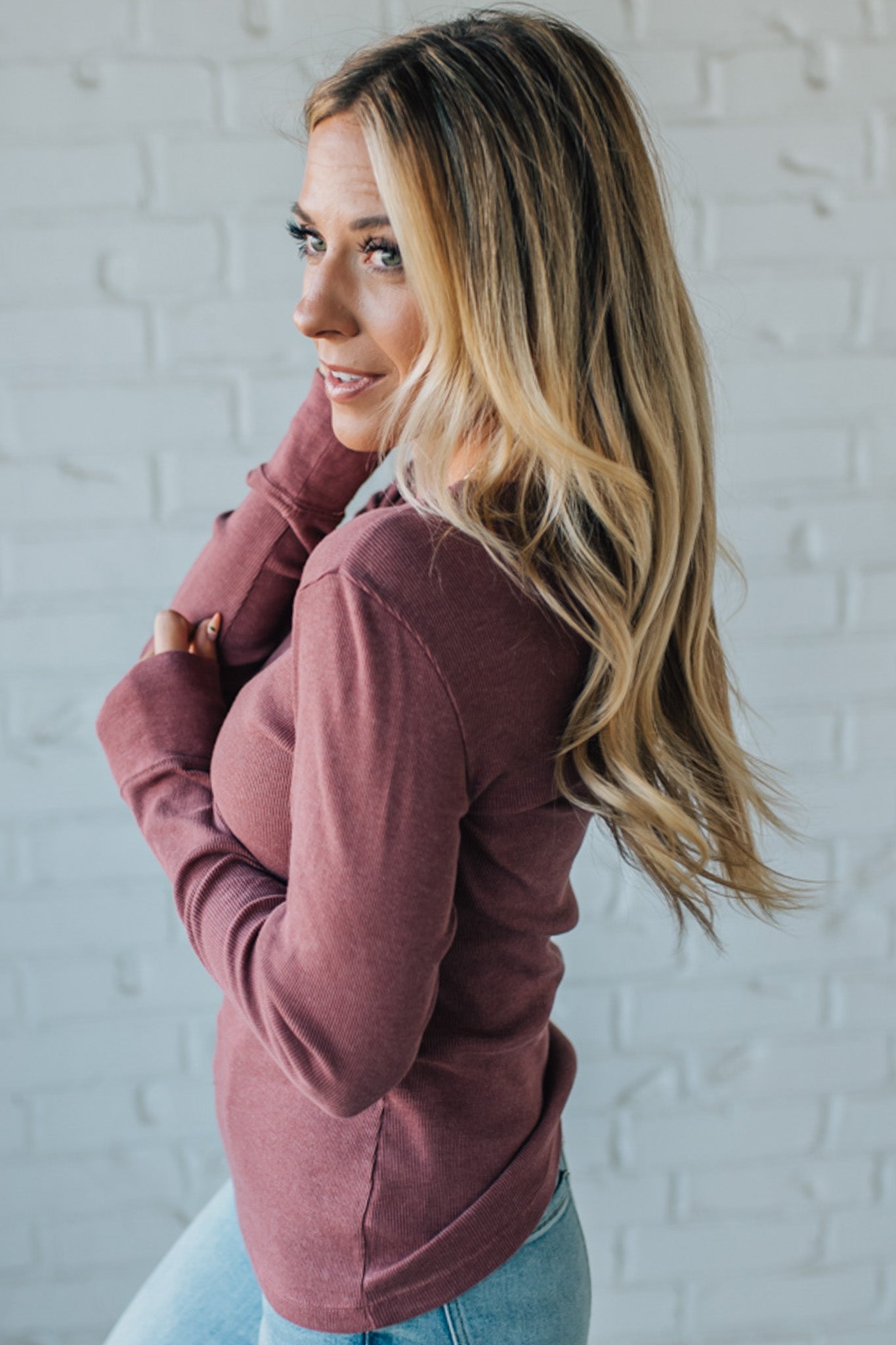 Woman wearing a maroon hoodie and light blue jeans against a white brick wall.