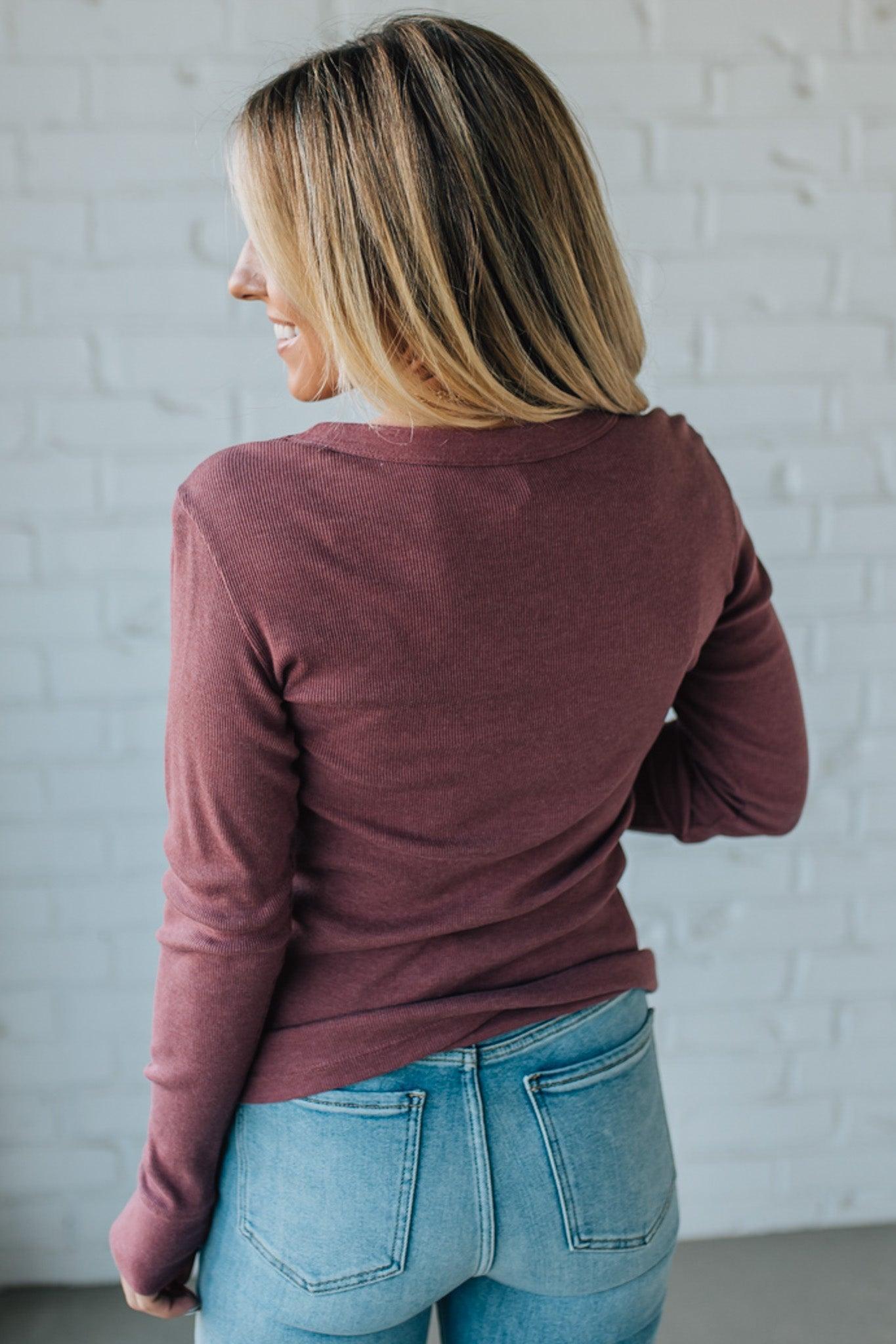 Person wearing a maroon long-sleeve top and blue jeans against a white brick wall.