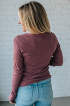 Person wearing a maroon long-sleeve top and blue jeans against a white brick wall.