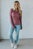 Woman wearing a mauve long-sleeve top and light blue jeans against a white brick wall.