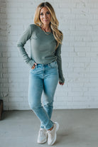 Woman wearing a green sweater and light blue jeans standing against a white brick wall.