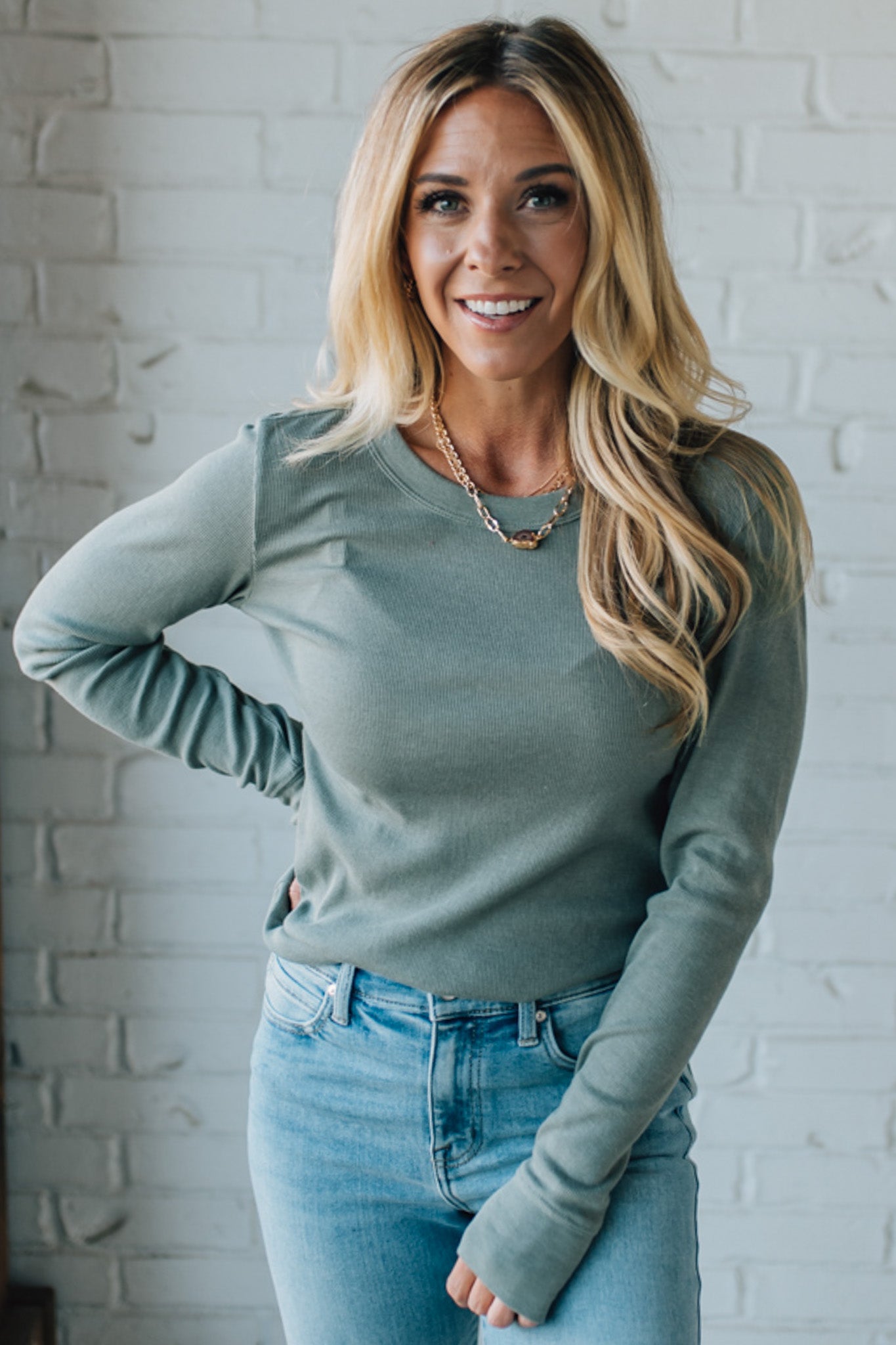 Woman wearing a green long-sleeve top and blue jeans against a white brick wall.