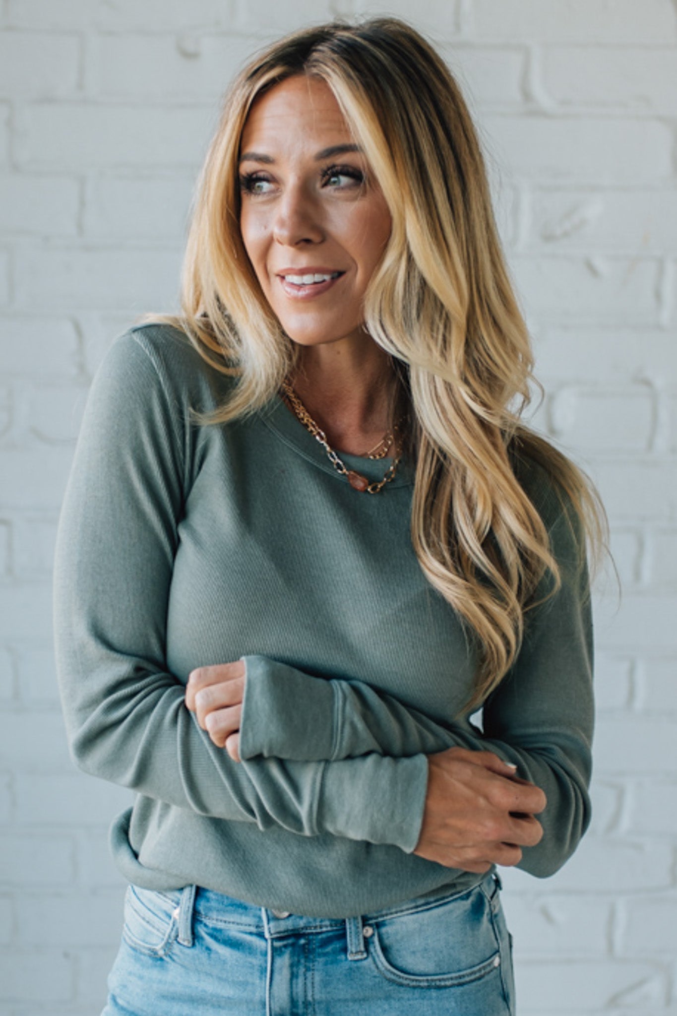 Woman wearing a green sweater and blue jeans against a white brick wall.