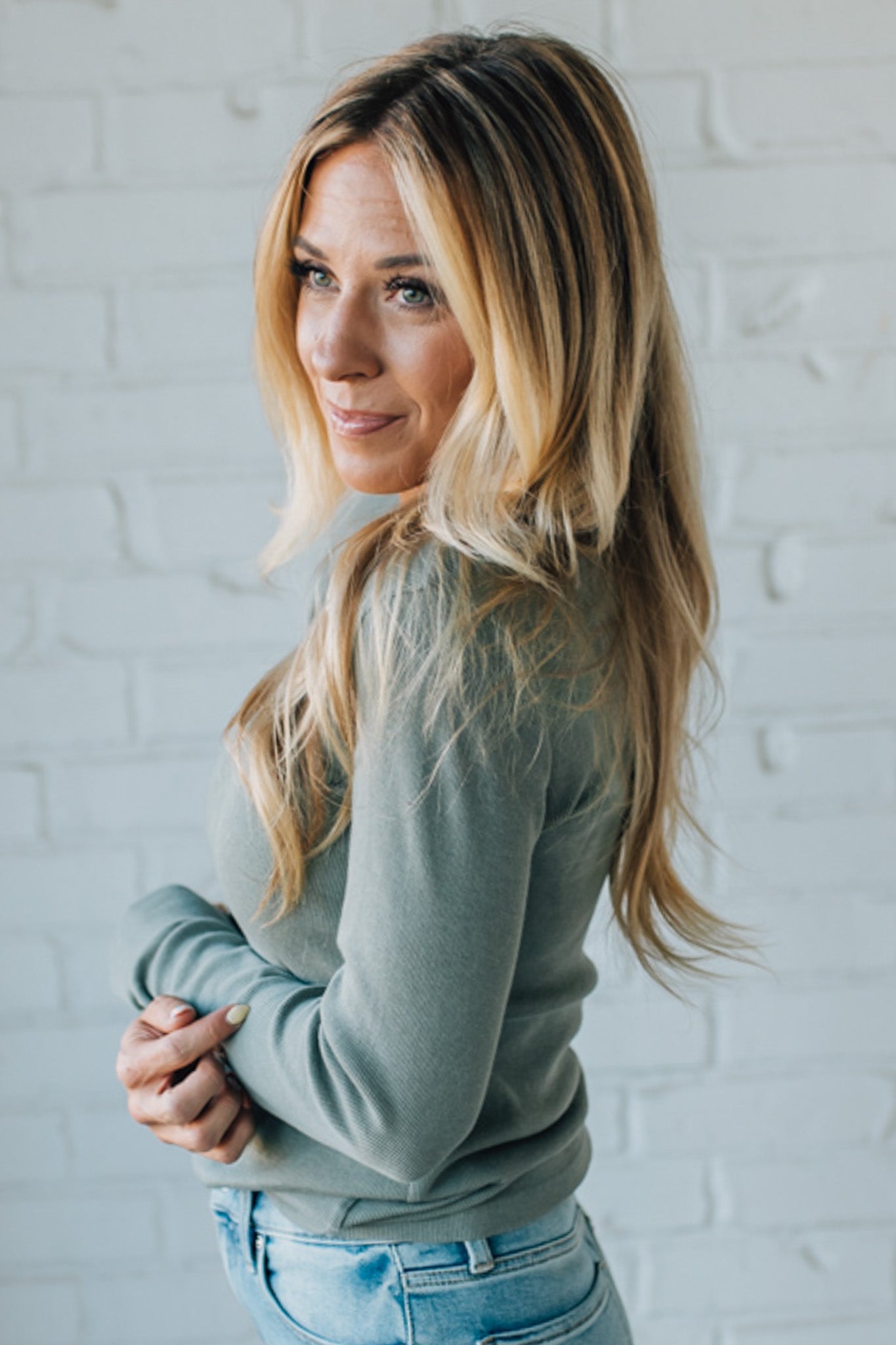 Woman wearing a light green sweater and blue jeans against a white brick wall.