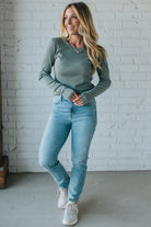 Woman wearing a green long-sleeve top and light blue jeans against a white brick wall.