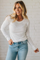 Woman wearing a white long-sleeve top and light blue jeans against a white brick wall.