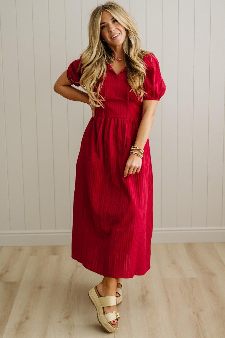 Woman wearing a red dress standing against a light wooden paneled wall.