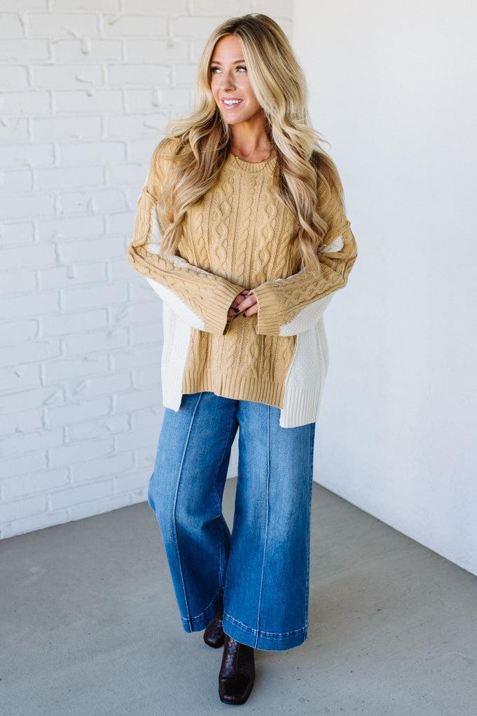 Woman wearing a beige cable knit sweater, white shirt, blue jeans, and black shoes against a white brick wall.