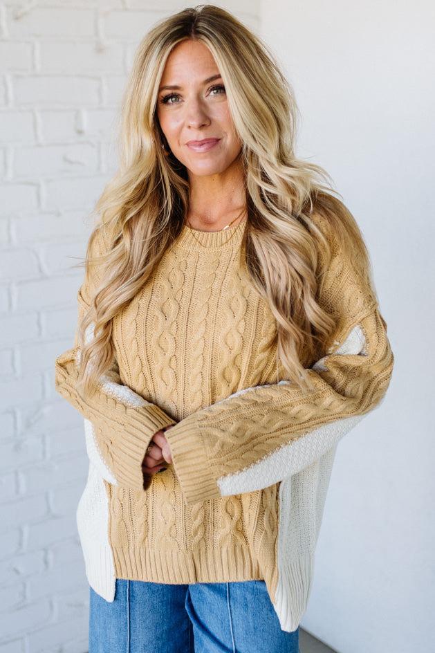 Woman wearing a beige cable knit sweater with white accents against a white brick wall.