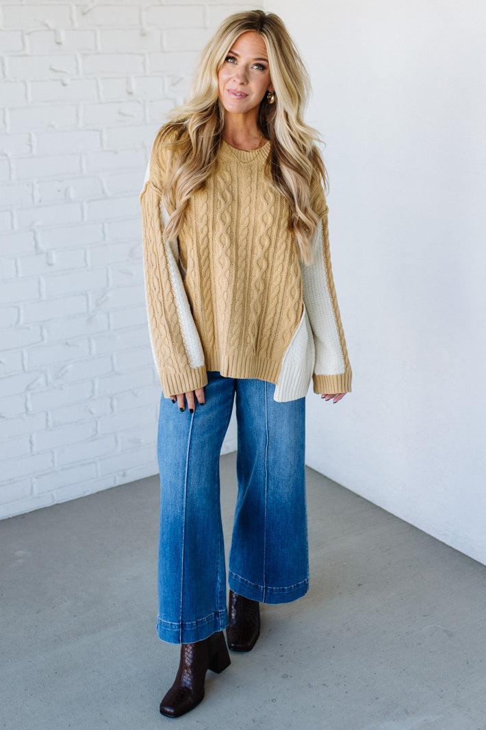 Woman wearing a beige sweater with white sleeves, blue jeans, and brown boots against a white brick wall.