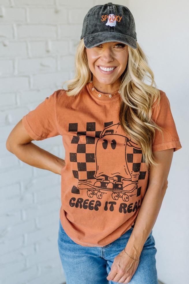 Woman wearing a brown t-shirt with a graphic design and a cap, standing against a white brick wall.