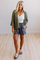 Woman wearing a green cardigan, white top, blue shorts, and sandals indoors.