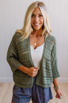Woman wearing a green patterned cardigan over a white top and blue shorts, standing against a plain background.