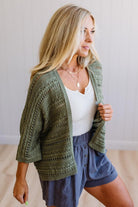 Woman wearing a green knitted cardigan over a white top and blue shorts indoors.