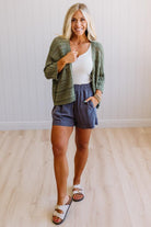 Woman wearing a green cardigan, white top, and blue shorts indoors.