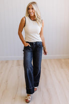 Woman wearing a white sleeveless top and blue jeans standing in a room with a light wooden floor and white walls.