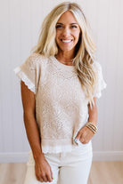 A smiling blonde woman wearing a cream-colored, short-sleeve open-knit top with ruffled sleeves and white pants, standing against a white paneled wall.