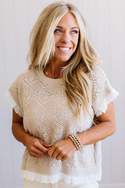 A smiling blonde woman wearing a cream-colored, short-sleeve knit top with ruffled sleeves and a ruffled hem, paired with white pants and gold bracelets.