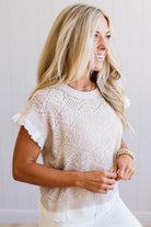 A woman wearing a cream-colored, short-sleeve knit top with ruffle trim and a textured openwork pattern, paired with white pants against a light neutral wall.