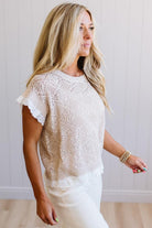 A woman wearing a cream-colored, short-sleeve open-knit sweater with delicate ruffle trim on the sleeves and hem, paired with white pants, standing against a white paneled wall.