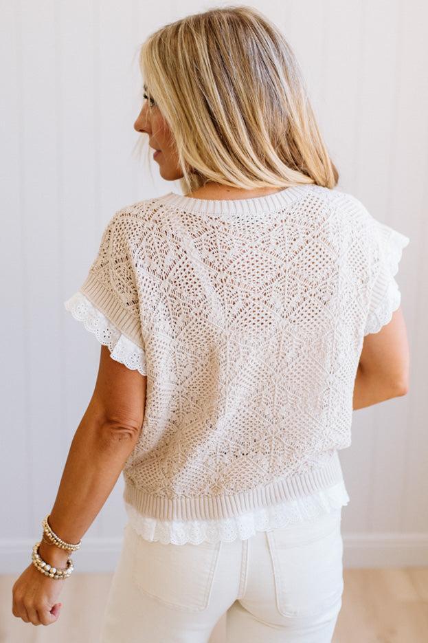 A woman shown from the back wearing a white crochet knit top with ruffled short sleeves and white pants, standing against a light indoor wall.