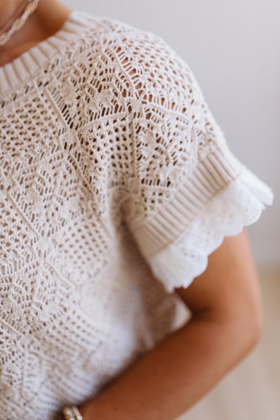 Close-up of a person wearing a cream-colored crochet knit top with short ruffled sleeves and an intricate openwork pattern.