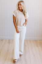 A woman modeling a cream-colored short-sleeve knit top with ruffled sleeves and matching cream wide-leg pants, standing in a brightly lit room with light wood floors and white paneled walls.