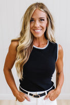 A smiling blonde woman wearing a black sleeveless knit top with white trim along the neckline and armholes, paired with white pants and a black belt, against a light neutral indoor background.