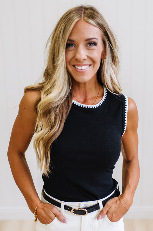 A smiling blonde woman wearing a black sleeveless knit top with white trim along the neckline and armholes, paired with white pants and a black belt, against a light neutral indoor background.