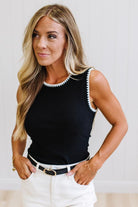 A woman modeling a black sleeveless knit top with white scalloped trim along the neckline and armholes, paired with white pants and a black belt, against a light neutral indoor background.