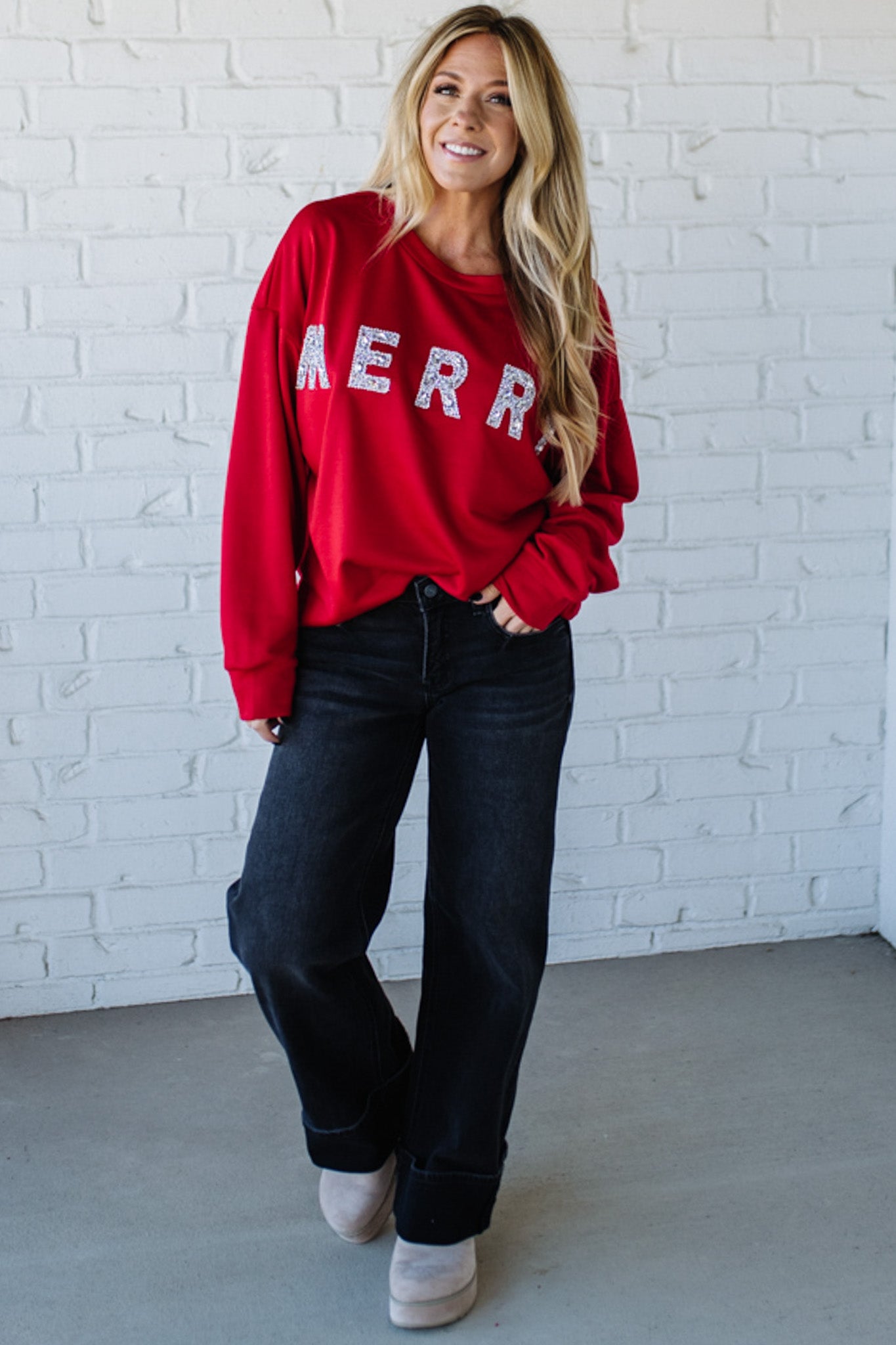 Woman wearing a red sweatshirt with 'MERRY' sparkled text against a white brick wall.