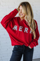 Woman wearing a red sweatshirt with 'MERRY' sparkled text against a white brick wall.