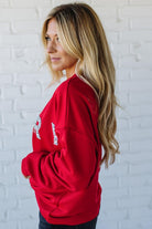 Woman wearing a red sweatshirt with 'MERRY' sparkled text against a white brick wall.