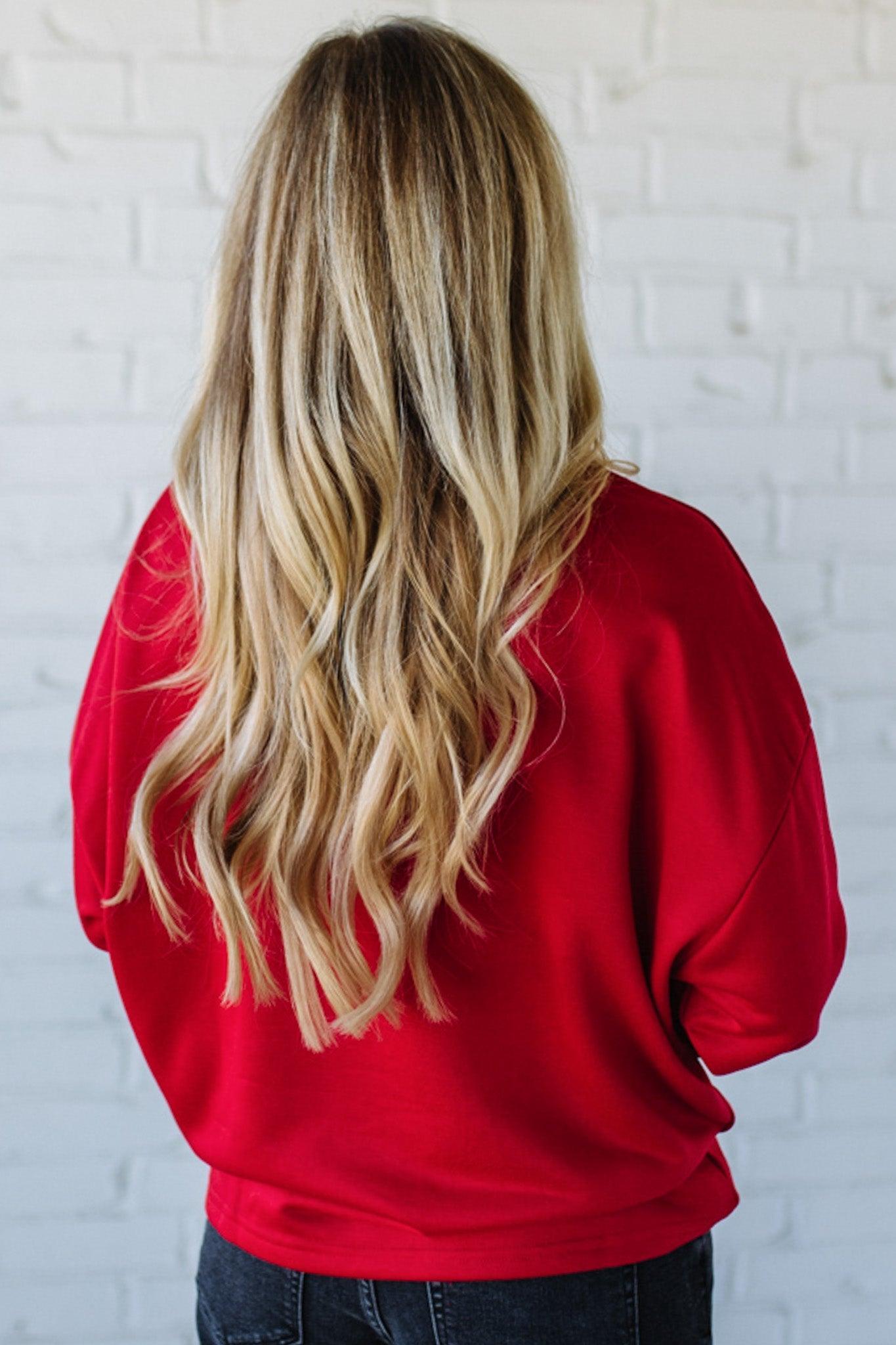 Woman wearing a red sweatshirt with 'MERRY' sparkled text against a white brick wall.