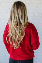 Woman wearing a red sweatshirt with 'MERRY' sparkled text against a white brick wall.