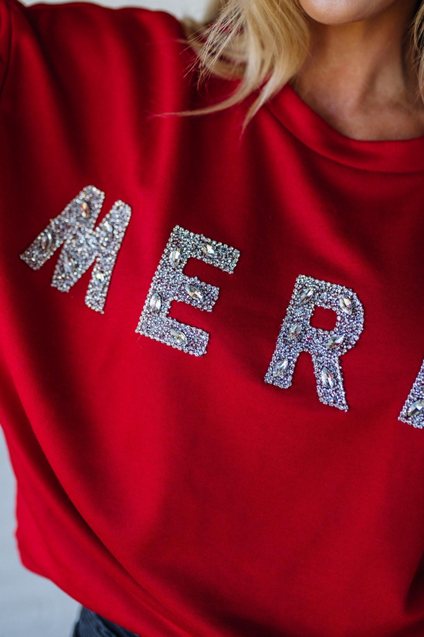 Woman wearing a red sweatshirt with 'MERRY' sparkled text against a white brick wall.