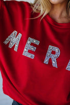 Woman wearing a red sweatshirt with 'MERRY' sparkled text against a white brick wall.