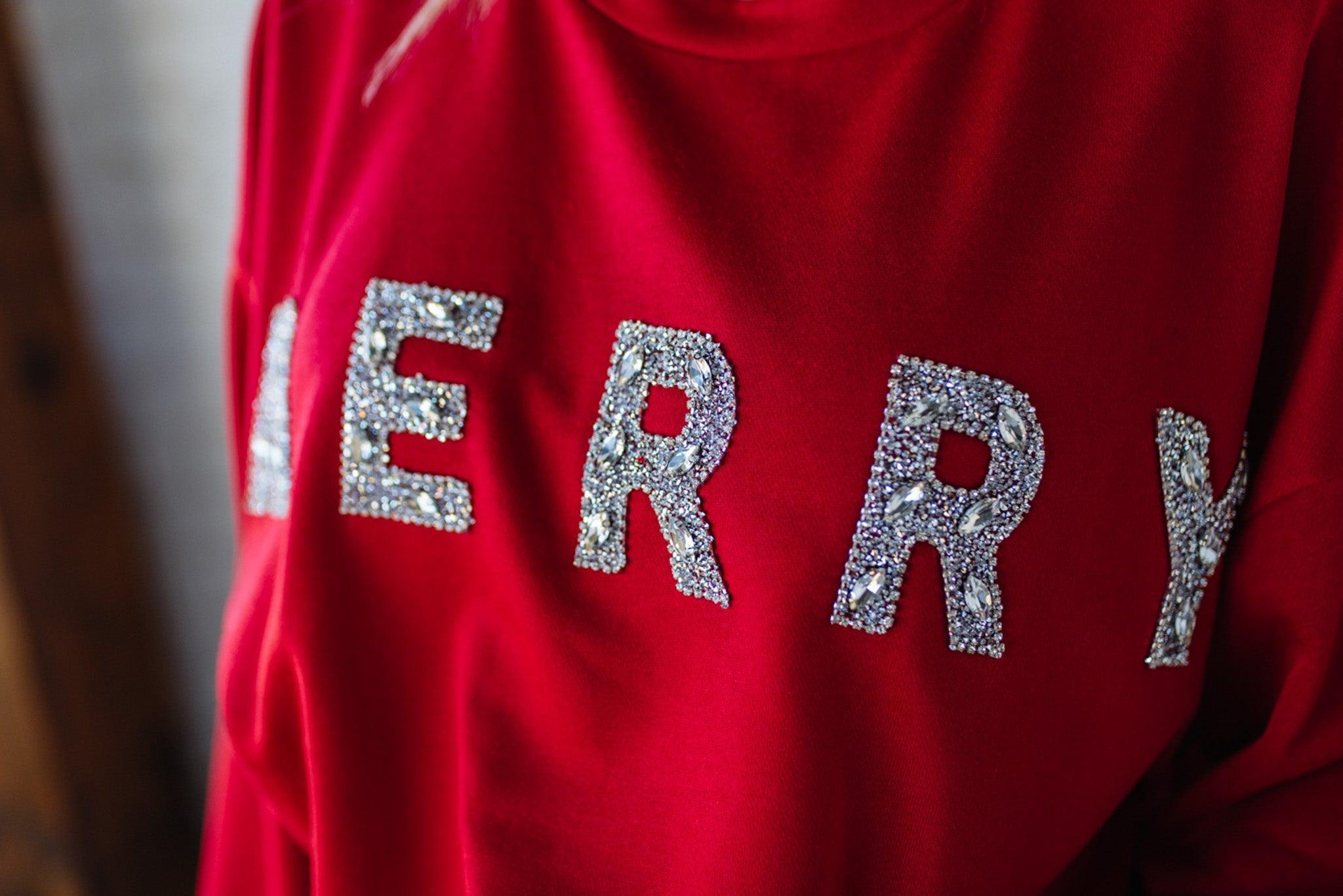 Woman wearing a red sweatshirt with 'MERRY' sparkled text against a white brick wall.