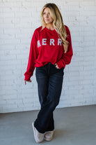Woman wearing a red sweatshirt with 'MERRY' sparkled text against a white brick wall.