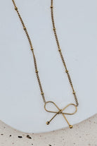 A gold necklace with a delicate beaded chain and a bow-shaped pendant, displayed on a white ceramic plate over a light beige textured surface.