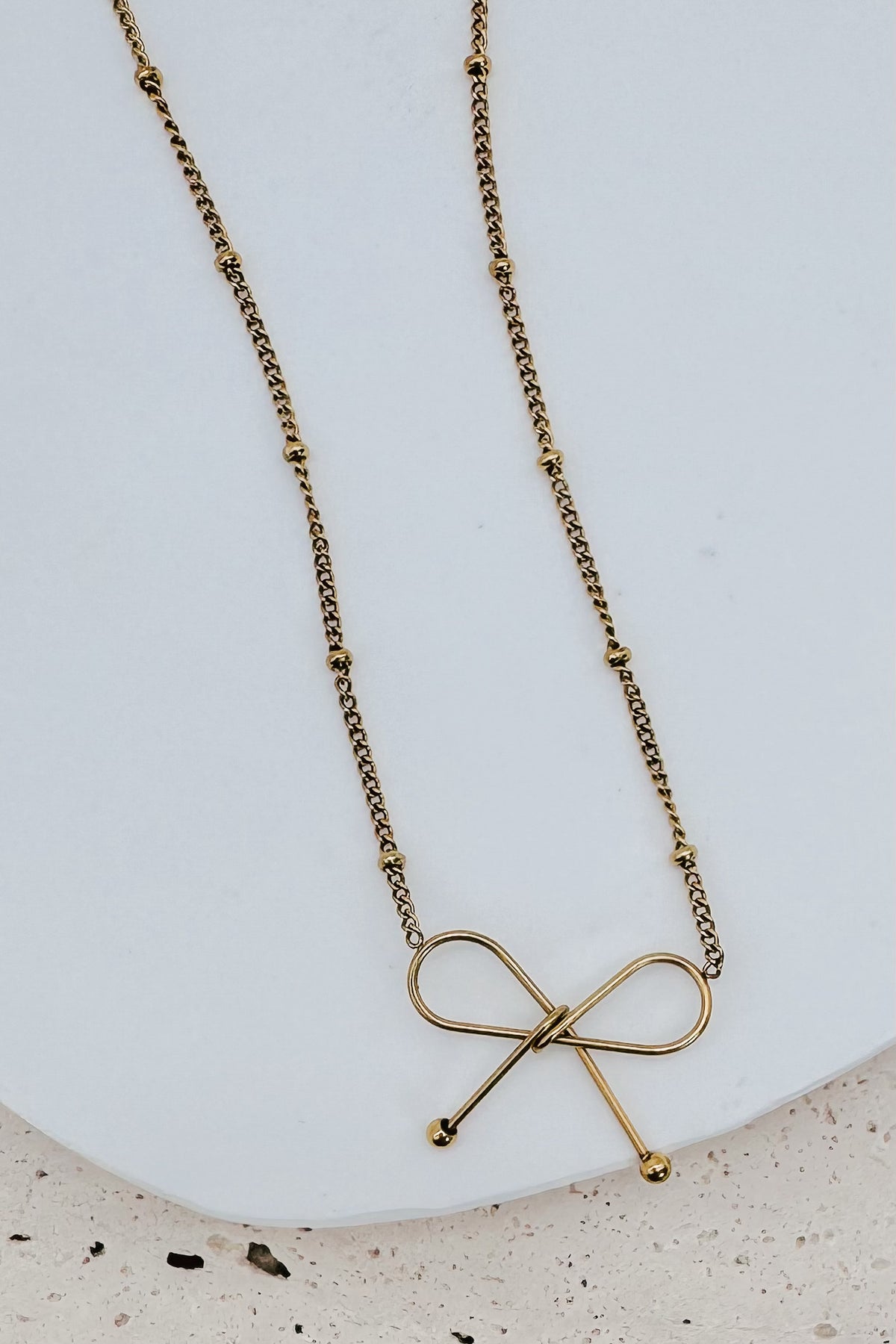 A gold necklace with a delicate beaded chain and a bow-shaped pendant, displayed on a white ceramic plate over a light beige textured surface.