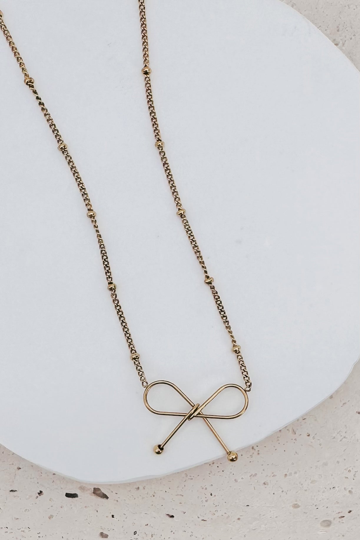 A delicate gold chain necklace with a minimalist bow-shaped pendant, displayed on a white ceramic plate over a light beige textured surface.