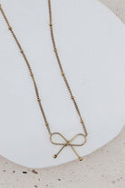 A delicate gold chain necklace with a minimalist bow-shaped pendant, displayed on a white ceramic plate over a light beige textured surface.