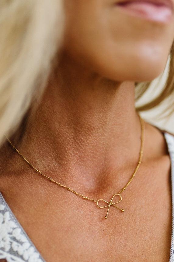 A close-up lifestyle shot of a woman wearing a delicate gold chain necklace with a small bow-shaped pendant.