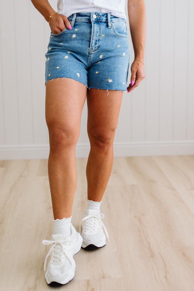 Person wearing blue denim shorts with white flowers and white sneakers on a light wooden floor.