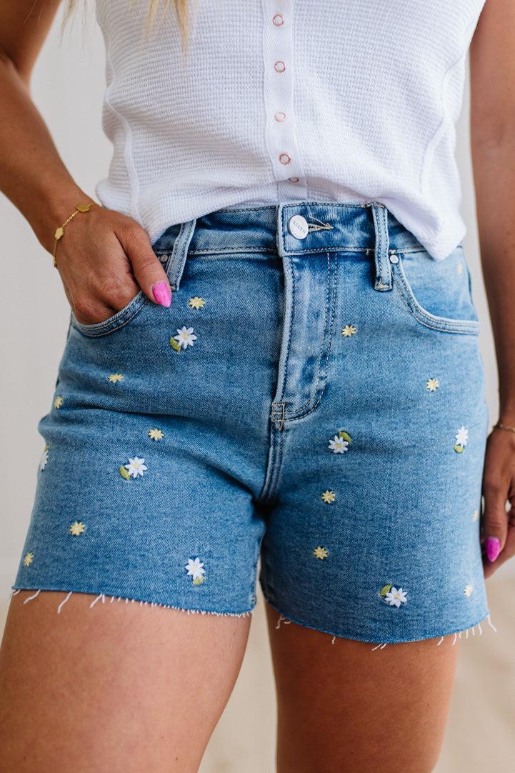 Blue denim shorts with floral embroidery worn by a person against a neutral background