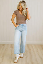 Woman wearing a brown top and light blue jeans standing in a room with wooden flooring and a white wall.